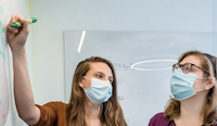 Two white women wearing surgical masks looking at a white board