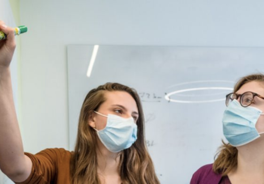 Two white women wearing surgical masks looking at a white board